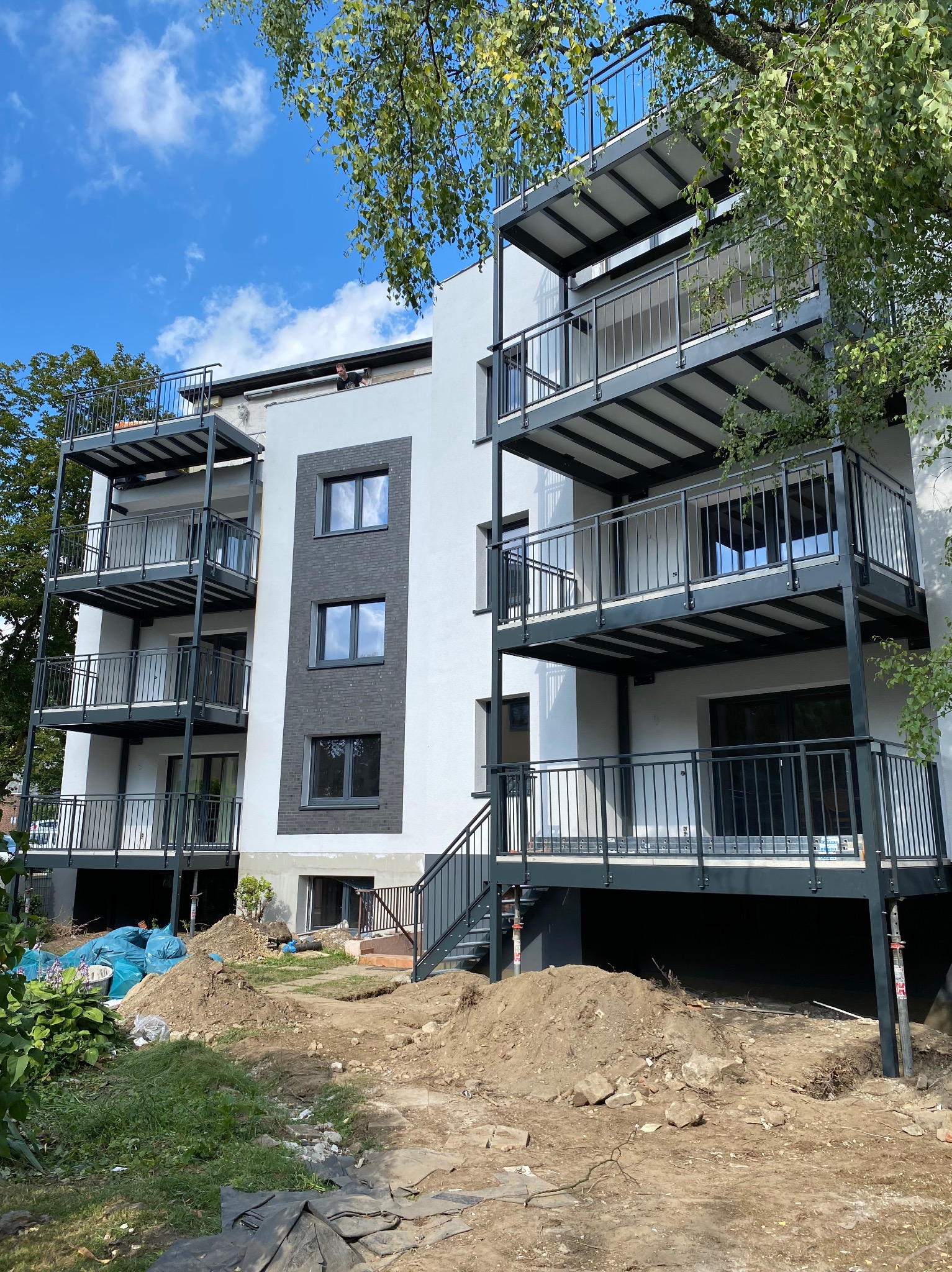 Wielopiętrowy budynek z balkonami, częściowo otynkowany na biało, z szarymi akcentami. Teren wokół budynku w trakcie zagospodarowywania.