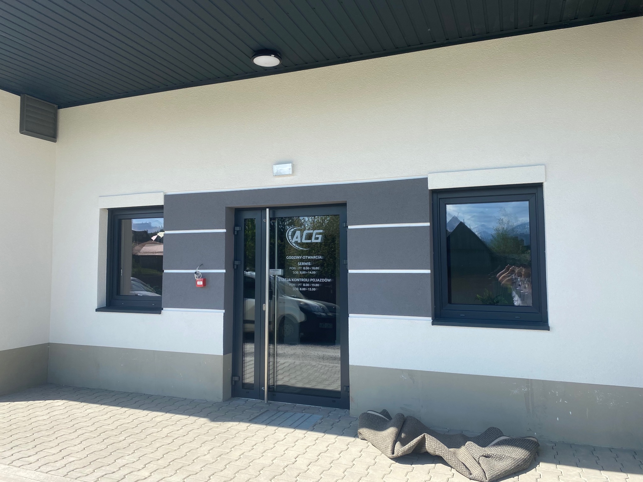Biała elewacja budynku z szarą lamperią, ciemnymi oknami i drzwiami wejściowymi. Widoczny fragment chodnika z kostki brukowej i dywanik przed wejściem.