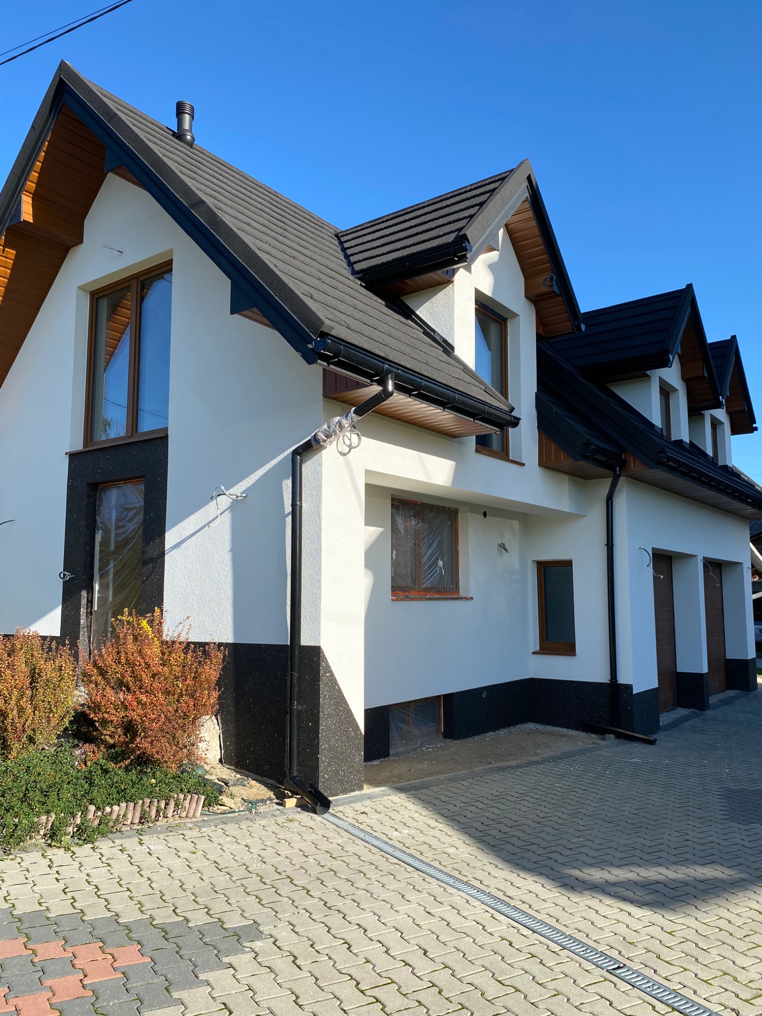Dwukondygnacyjny dom z białą elewacją i czarnymi akcentami, brązowymi oknami. Część okien zabezpieczona folią. Kostka brukowa przed budynkiem.