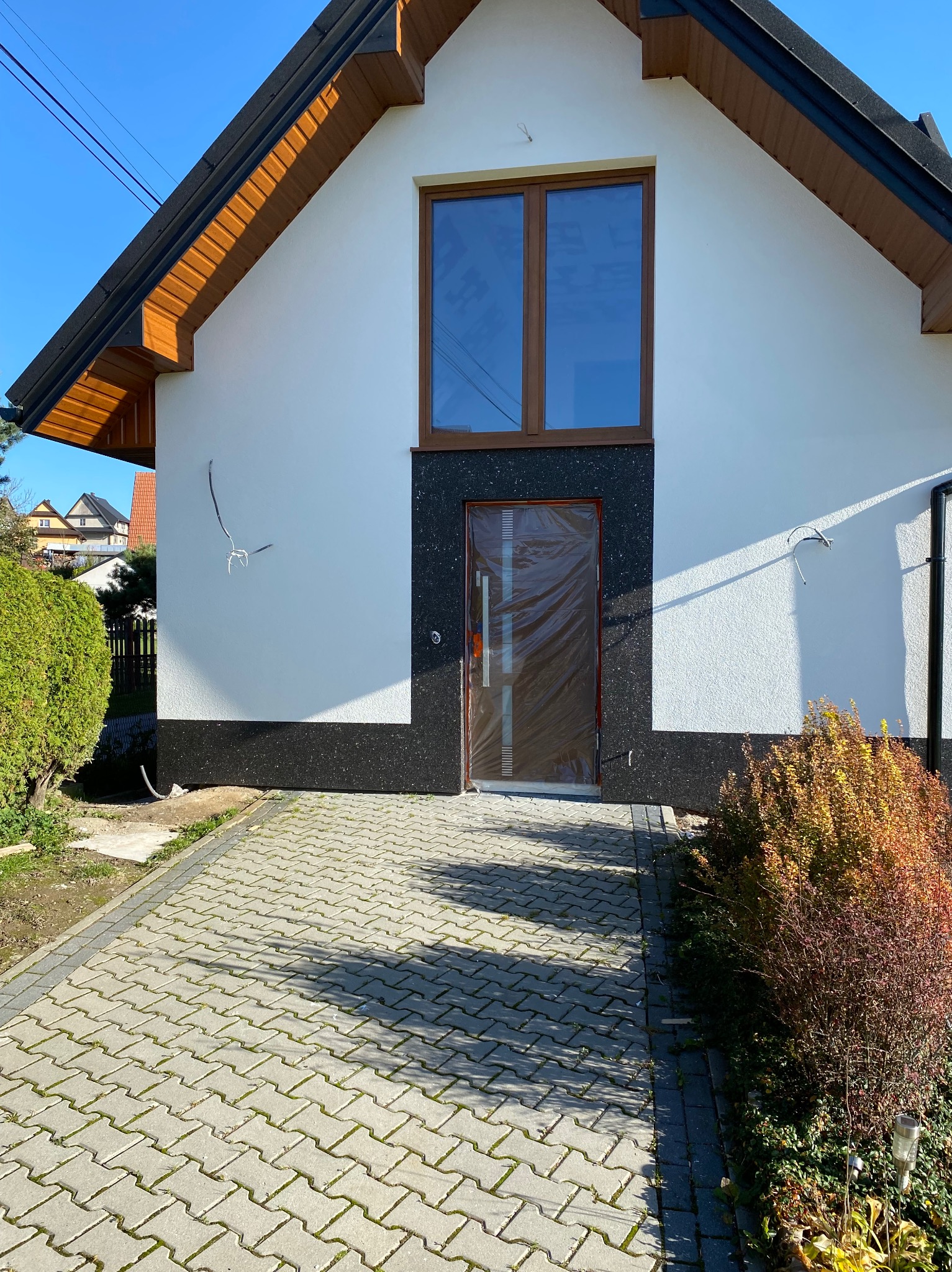 Wykończona elewacja domu jednorodzinnego z nową stolarką okienną i drzwiami wejściowymi zabezpieczonymi folią ochronną. Widoczne przewody na ścianie.