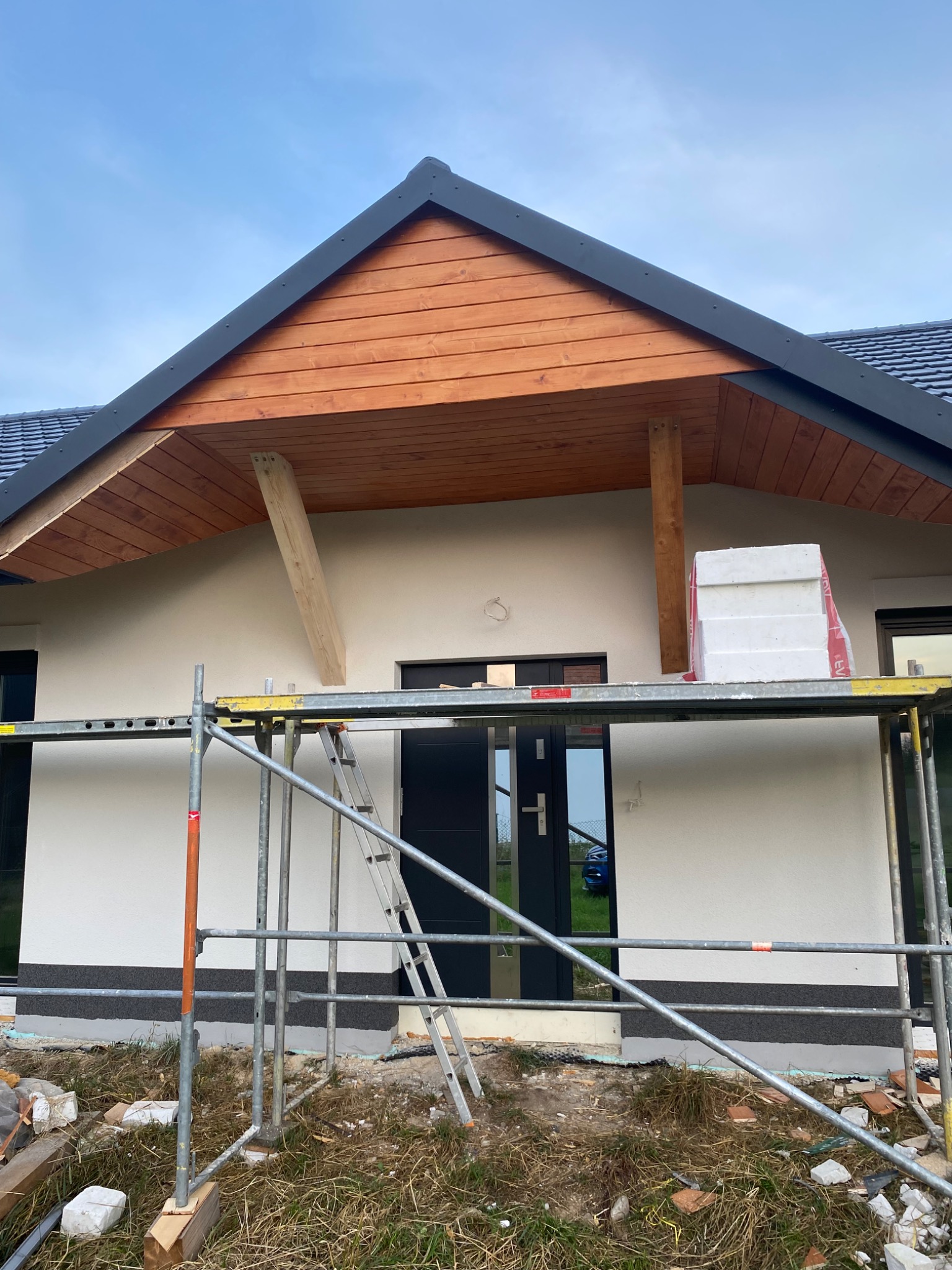 Dom jednorodzinny w trakcie prac elewacyjnych. Widoczne rusztowanie, styropian i fragment drewnianej konstrukcji dachu. Świeżo otynkowana ściana.