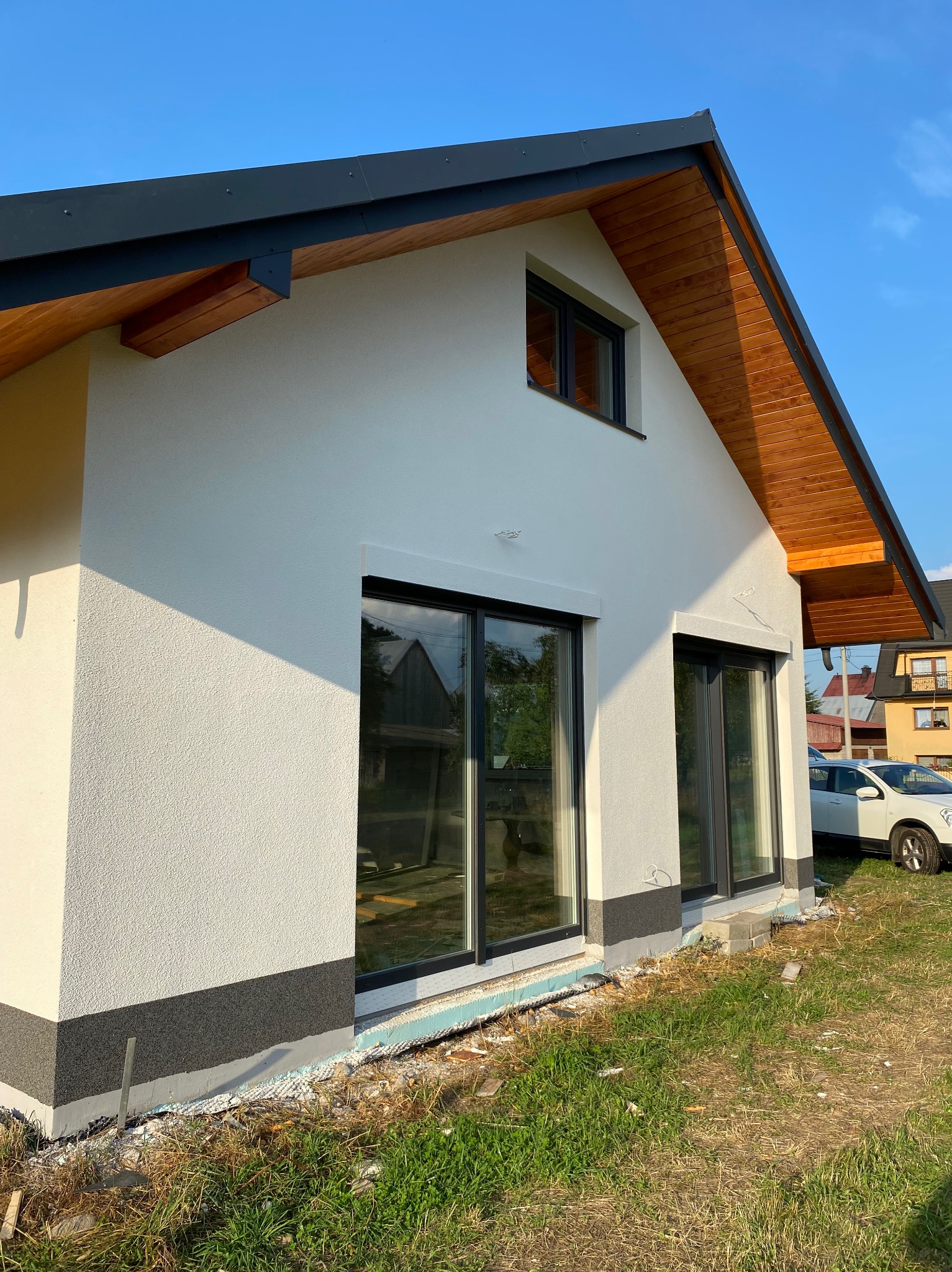 Biała elewacja domu jednorodzinnego z drewnianym podbitką dachu i czarnymi ramami okiennymi. Widoczny fragment ocieplenia przy gruncie i trawnik.