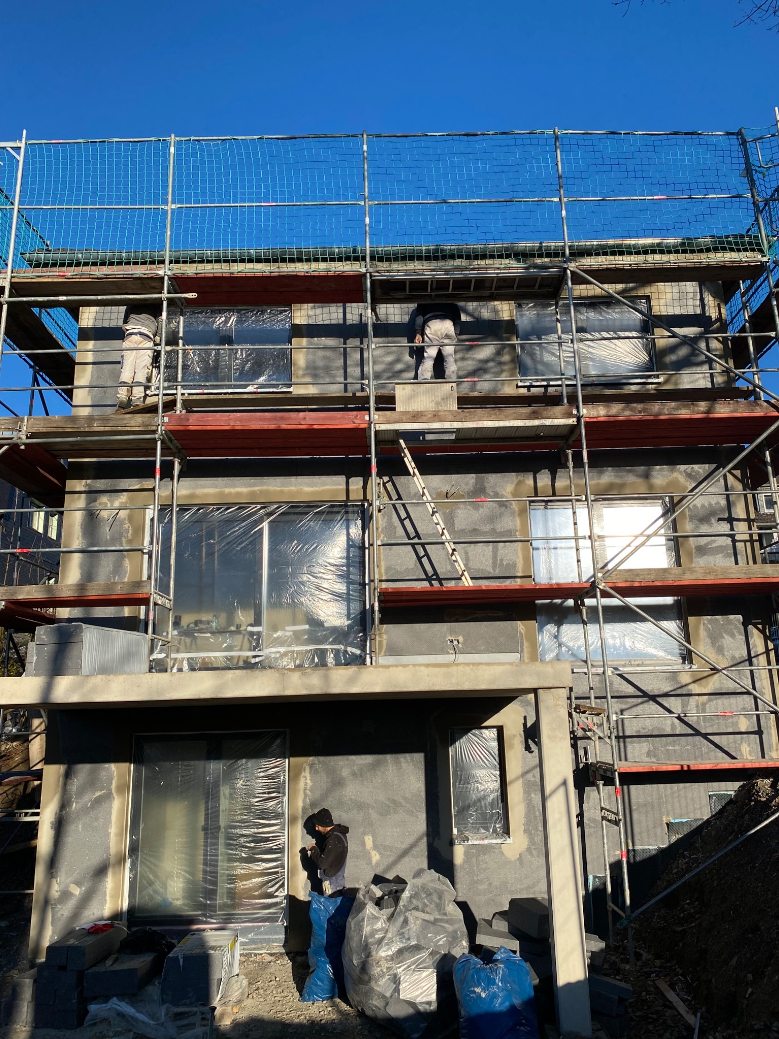 Dom w trakcie ocieplania: rusztowania, pracownicy, zabezpieczone okna folią. Widoczne materiały izolacyjne i przygotowana elewacja pod tynk. Słoneczny dzień.