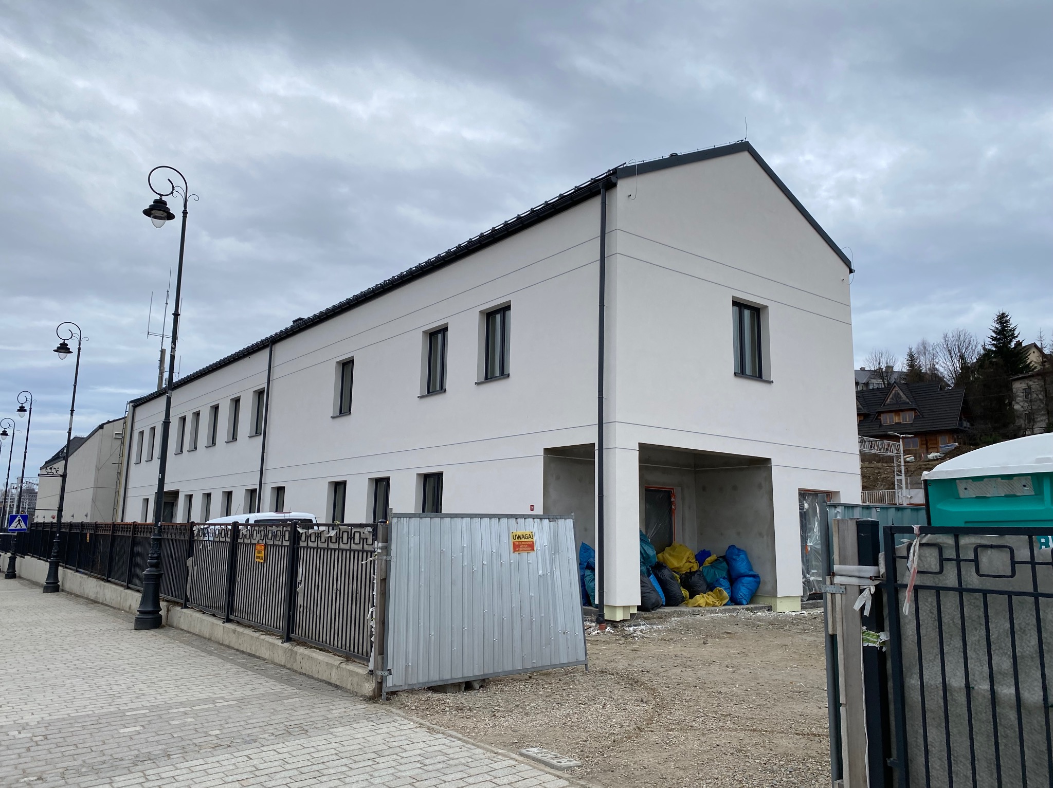 Nowoczesny budynek z białą elewacją w trakcie prac wykończeniowych, widoczny z chodnika. Uwagę zwracają czarne ramy okienne i minimalistyczny styl.