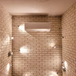 Biała jednostka wewnętrzna klimatyzatora zamontowana na ścianie z cegły, oświetlona przez cztery małe reflektory punktowe, z widocznymi przewodami elektrycznymi.