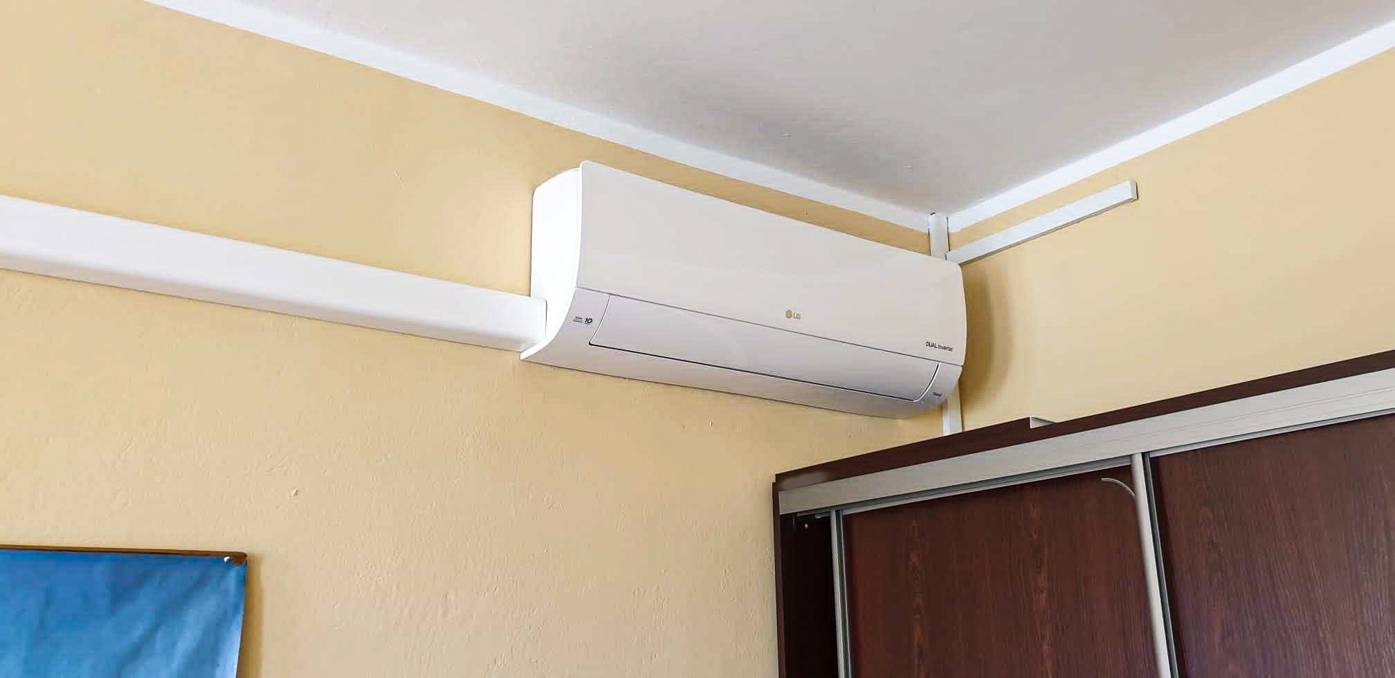 Biały klimatyzator LG zamontowany na beżowej ścianie nad brązową szafą przesuwną w pokoju.