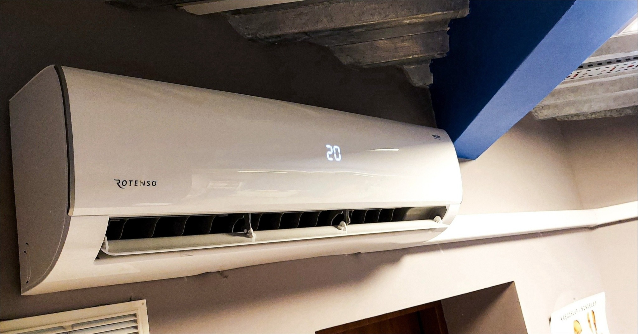 Biała jednostka klimatyzacji Rotenso z wyświetlaczem temperatury 20 stopni, zamontowana na szarej ścianie pod betonowym sufitem z niebieskim elementem wykończeniowym.