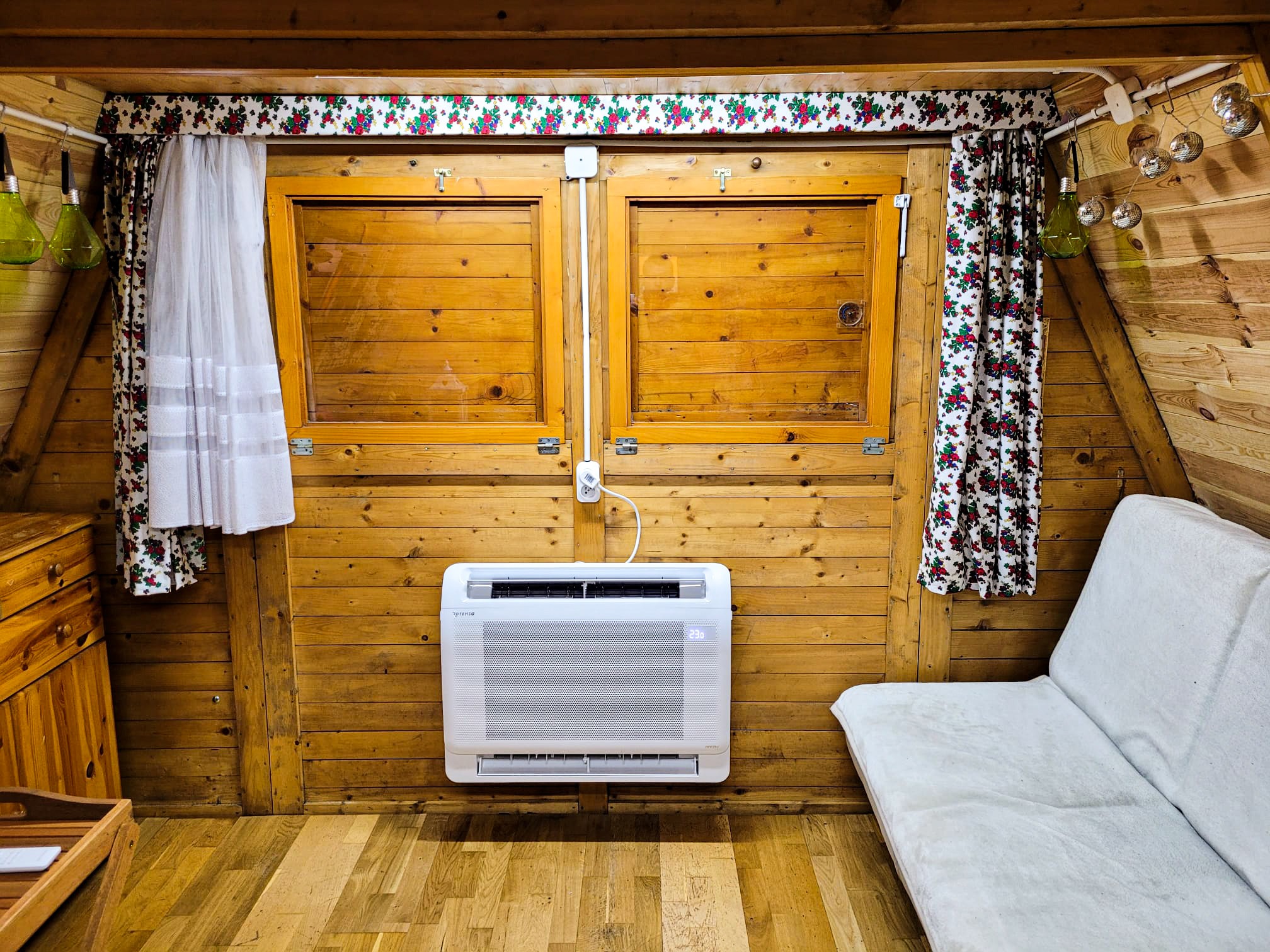Klimatyzator zamontowany w drewnianym pokoju na poddaszu, z widocznym drewnianym wykończeniem ścian, zasłonami w kwiaty i elementami dekoracyjnymi.
