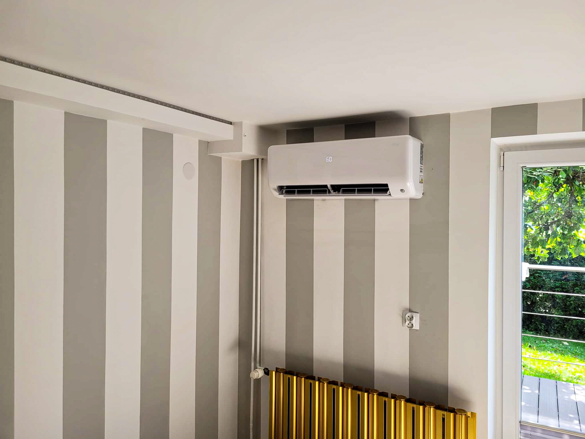 Biała klimatyzacja ścienna z wyświetlaczem 60, zamontowana na ścianie w szaro-białe pasy, z widocznym złotym grzejnikiem żeberkowym i oknem z widokiem na zieleń.