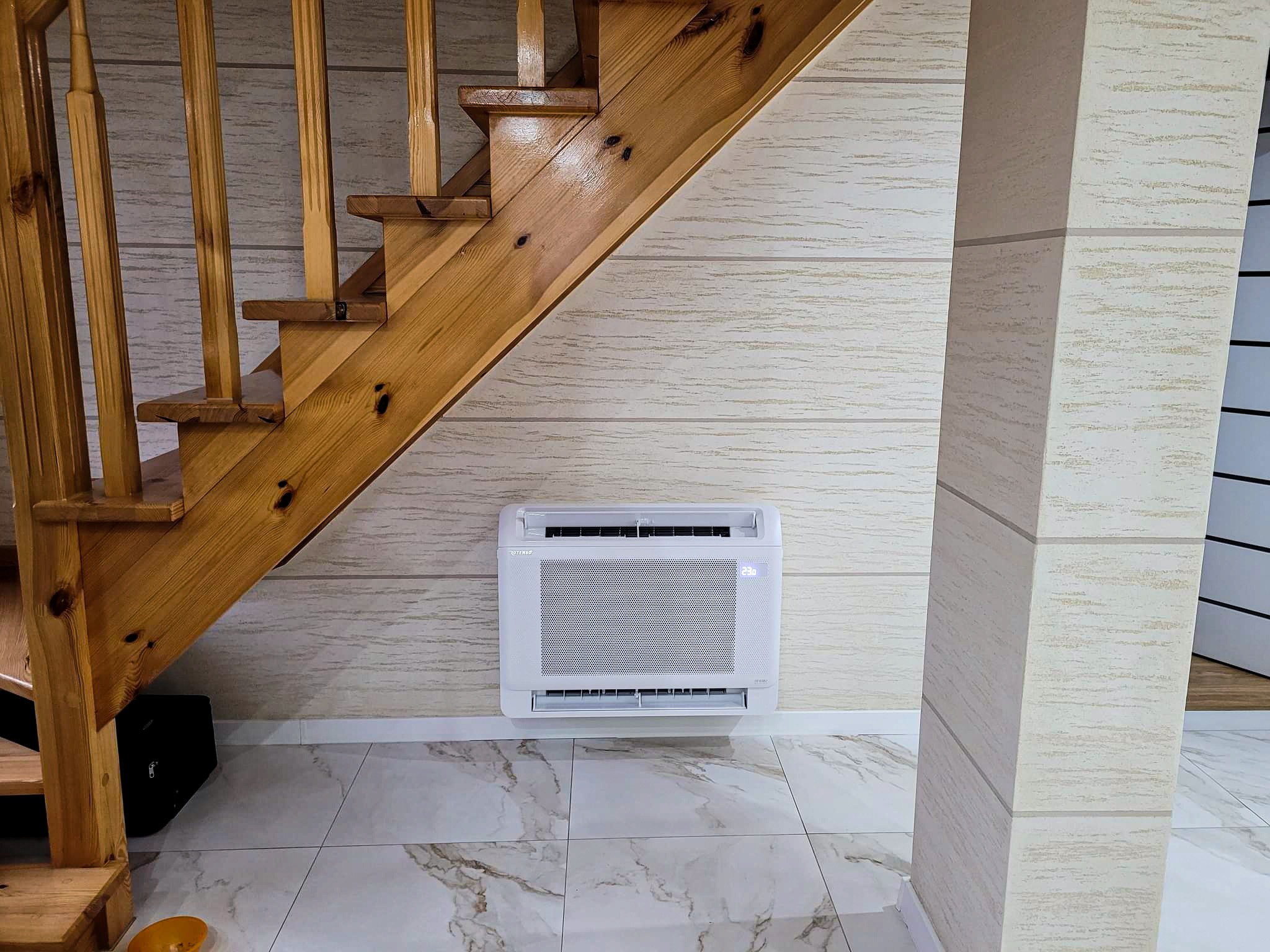 Biała jednostka klimatyzacyjna zamontowana na ścianie pod drewnianymi schodami w domu, widoczny wyświetlacz z temperaturą 23 stopnie Celsjusza.