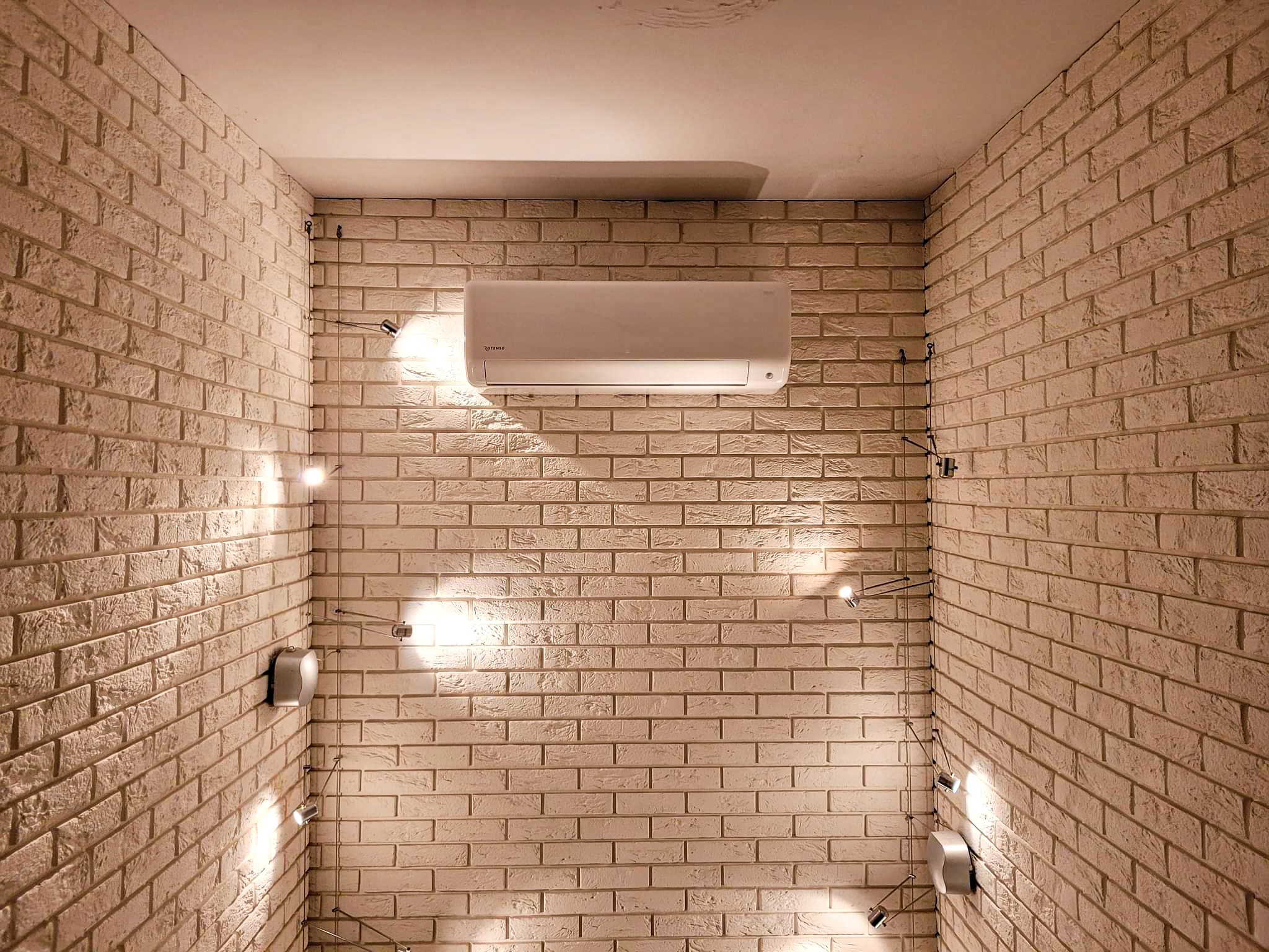 Biała jednostka wewnętrzna klimatyzatora zamontowana na ścianie z cegły, oświetlona przez cztery małe reflektory punktowe, z widocznymi przewodami elektrycznymi.