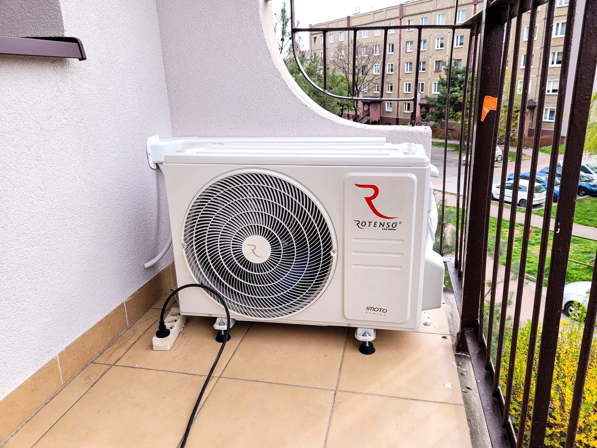 Biała jednostka zewnętrzna klimatyzatora Rotenso zamontowana na balkonie mieszkania w bloku, widok z boku na kratkę wentylacyjną.