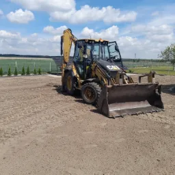 Żółta koparko-ładowarka CAT na ubitym gruncie, widoczne ślady opon, w tle rząd tuj, pole uprawne i błękitne niebo z chmurami.