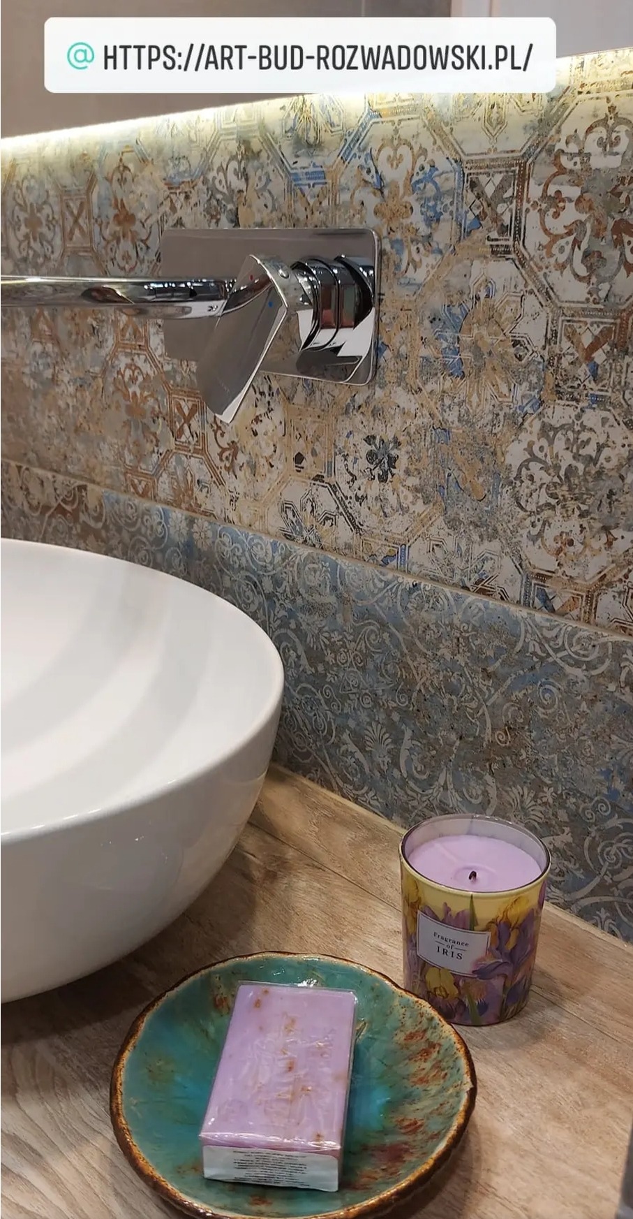 Umywalka nablatowa z chromowaną baterią i dekoracyjnymi, wzorzystymi płytkami w łazience, na blacie ceramiczna mydelniczka z fioletowym mydłem i zapalona świeczka o zapachu irysów.