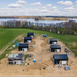 Widok z lotu ptaka na plac budowy osiedla domków jednorodzinnych z czarnymi dachami, z widocznymi panelami na jednym z domów i elementami konstrukcyjnymi, na tle zielonych łąk i jeziora.