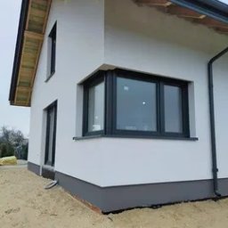 Sigmabud - Nowoczesny dom jednorodzinny z białymi ścianami i ciemnymi oknami narożnymi, w trakcie prac wykończeniowych elewacji, widoczne elementy konstrukcji dachu i piasek wokół budynku.