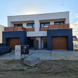 Bliźniak z płaskim dachem, wykończony w bieli i granacie, z drewnianymi bramami garażowymi i balustradami balkonów; na pierwszym planie stos kostki brukowej i zaparkowany samochód.