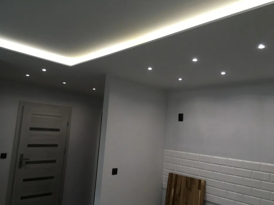 Nowoczesne oświetlenie sufitowe z listwą LED i punktowymi lampami w wyremontowanym wnętrzu z białymi ścianami i płytkami imitującymi cegłę.