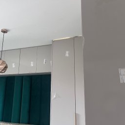 Mark Pol Meble - Montaż szafy wnękowej z szarej płyty MDF, widoczne folie ochronne, nad nią szafki wiszące i designerska lampa, zielony zagłówek łóżka w tle.