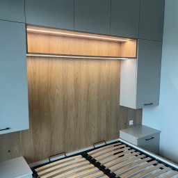 Mark Pol Meble - Szafa wnękowa z drewnianym panelem i podświetleniem LED nad łóżkiem. Szare szafki wiszące z czarnymi uchwytami, minimalistyczny design w sypialni.