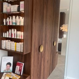 Mark Pol Meble - Szafa na wymiar z brązowego drewna, z półkami wypełnionymi kosmetykami w salonie kosmetycznym. Złote uchwyty i lustrzane wykończenie dodają elegancji.