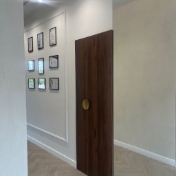 Mark Pol Meble - Nowoczesne wnętrze z drewnianymi drzwiami z okrągłą, złotą klamką, odbijającymi się w lustrze. Na ścianie galeria certyfikatów w czarnych ramkach i parkiet w jodełkę.