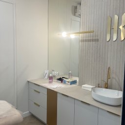 Mark Pol Meble - Minimalistyczna łazienka w Warszawie: białe szafki pod umywalką, złote akcenty, lustro i designerska lampa. Umywalka owalna, armatura złota.