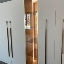 Mark Pol Meble - Biała szafa na wymiar z podświetlaną, przeszkloną witryną w złotej ramie. Minimalistyczne, długie uchwyty. Widok z bliska.