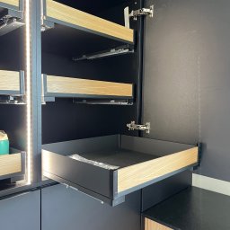 Mark Pol Meble - Ciemna szafa z wysuwanymi szufladami z jasnego drewna, widoczne prowadnice i okucia. Szuflady częściowo otwarte, wnętrze szafy oświetlone.