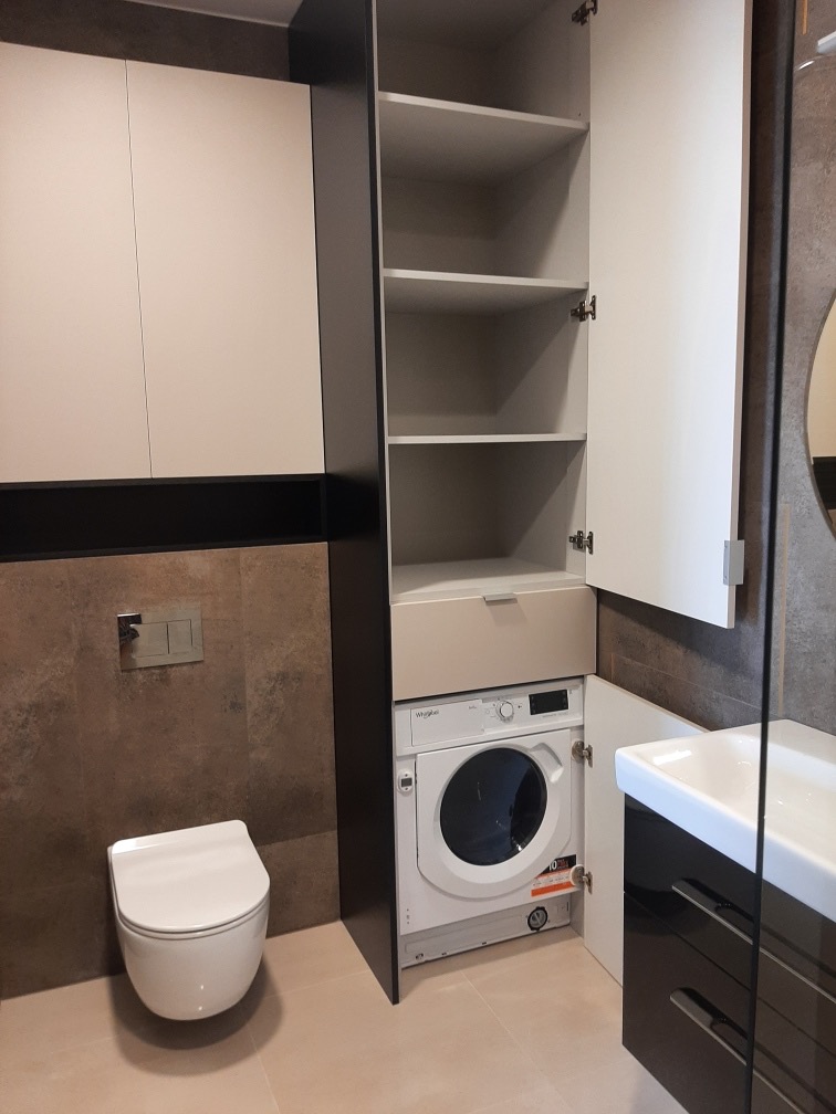 Nowoczesna łazienka z zabudową meblową: szafki wiszące, słupek z półkami, pralka w zabudowie, umywalka z szafką. Uwagę przykuwa spójna kolorystyka i funkcjonalne zagospodarowanie przestrzeni.