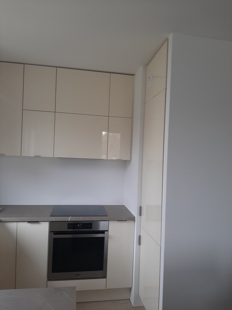 Jasna kuchnia z beżowymi, lakierowanymi frontami, szarym blatem i zabudowaną lodówką. Widoczna płyta indukcyjna i piekarnik Whirlpool.