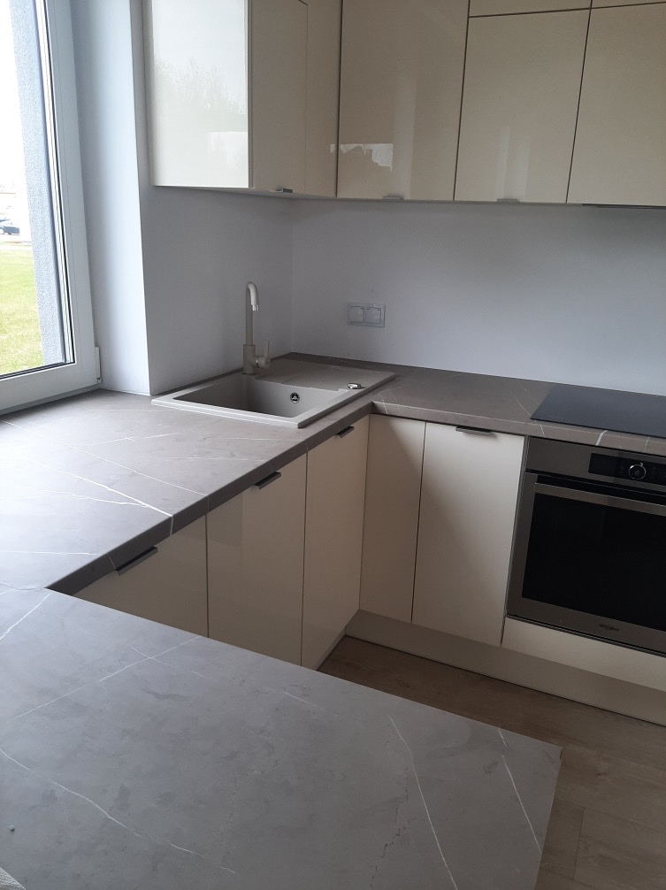 Jasna kuchnia z szarym blatem i zlewem przy oknie. Widoczny fragment płyty indukcyjnej i piekarnika. Górne szafki w kolorze kremowym. Minimalistyczny design.