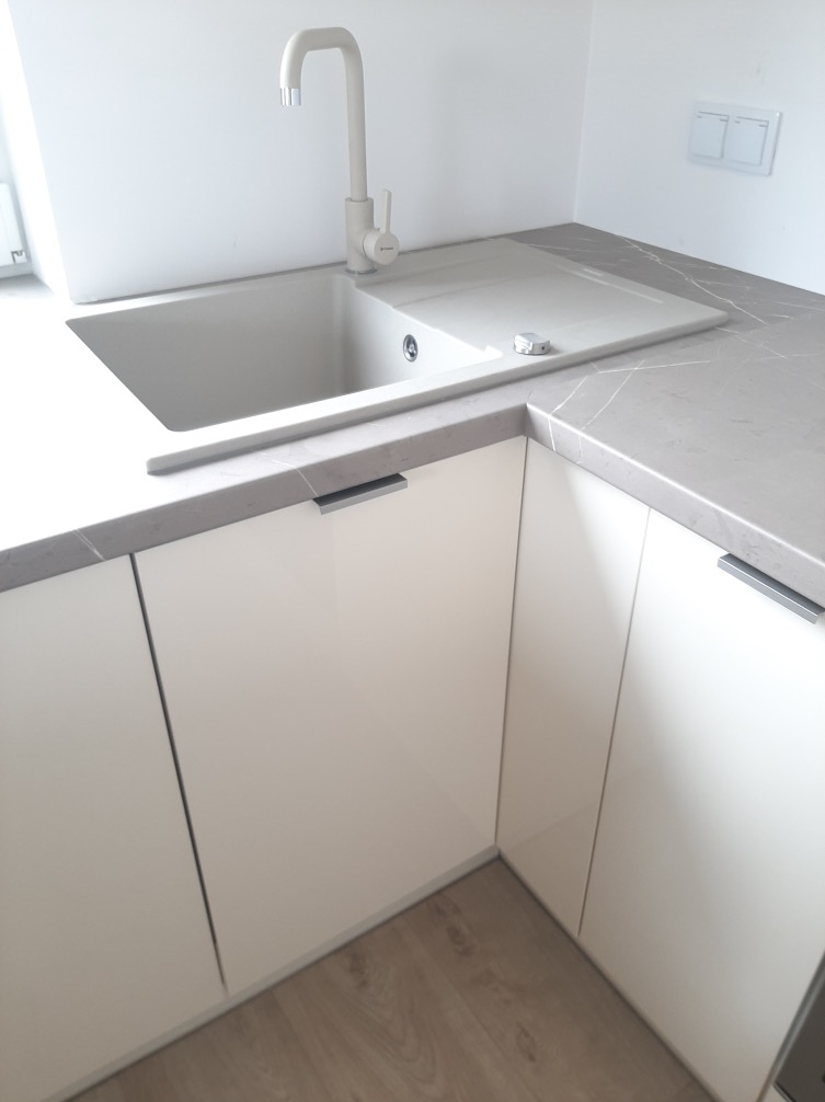 Biała, minimalistyczna kuchnia na wymiar: zlewozmywak z baterią, blat z szarym marmurkowym wzorem, gładkie fronty szafek z subtelnymi, podłużnymi uchwytami.