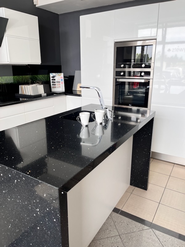 Błyszczący czarny blat z kropkami w nowoczesnej kuchni z białymi szafkami i srebrną armaturą. Widoczne dwie białe filiżanki i wbudowany piekarnik.