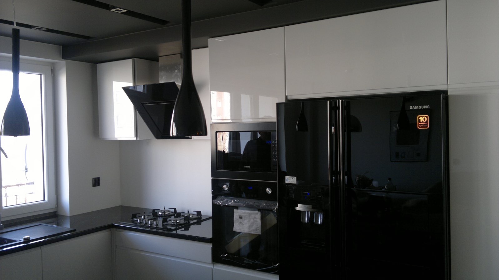 Nowoczesna kuchnia z białymi szafkami, czarnym blatem i sprzętem Samsung. Lampa wisząca nad kuchenką. Widok na okno i okap.
