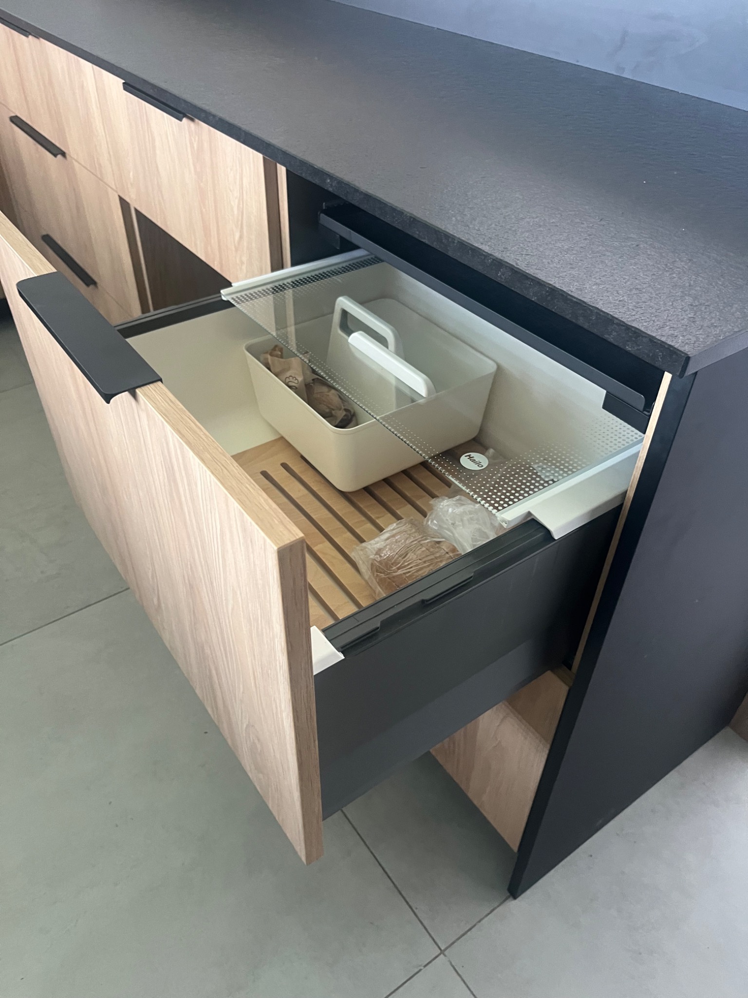 Otwarta szuflada z drewnianym wkładem i pojemnikami w szafce kuchennej z blatem w kolorze antracytu. Jasne drewno frontów i szary korpus.