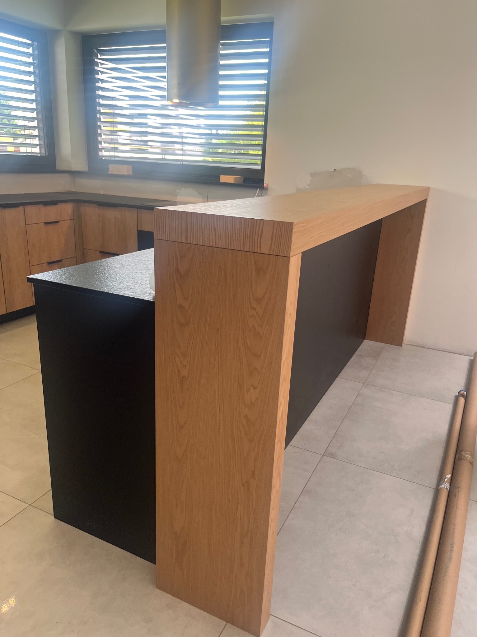 Wnętrze kuchni z wyspą o czarnym blacie i drewnianym wykończeniu. W tle zabudowa kuchenna i okno z żaluzjami. Widoczne rury na podłodze.