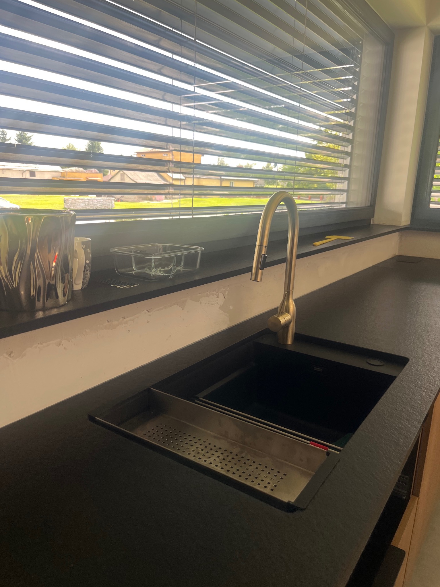 Ciemny blat kuchenny z wbudowanym zlewozmywakiem i złotą baterią, widok na okno z żaluzjami i zabudowę. Nowoczesny design z elementami wyposażenia.