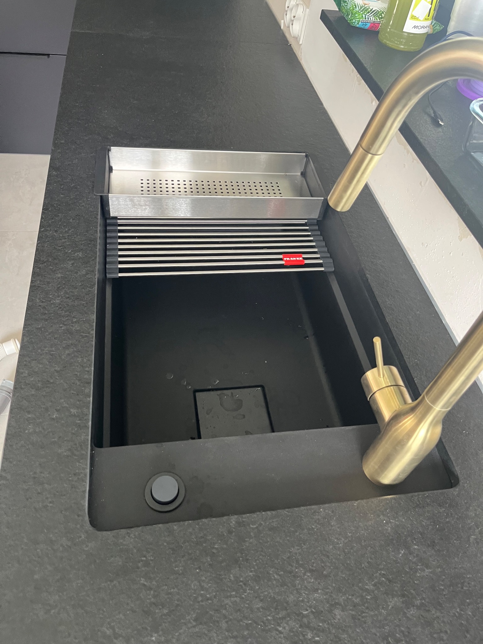 Czarny, granitowy blat kuchenny z wbudowanym, prostokątnym zlewem Franke i złotą baterią. Widoczny ociekacz i dozownik na płyn. Minimalistyczny design.