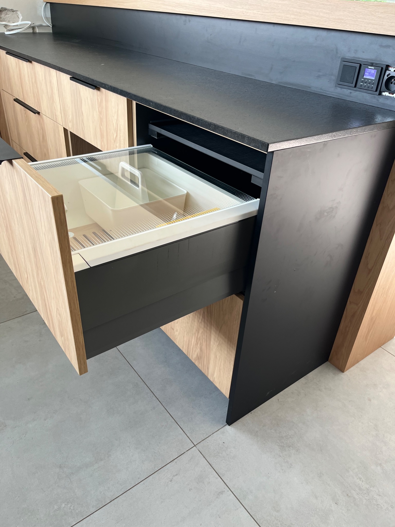 Nowoczesna kuchnia z blatem w kolorze antracytu i drewnianymi frontami. Wysuwana szuflada z organizerem na przybory kuchenne, widoczny detal wykończenia.