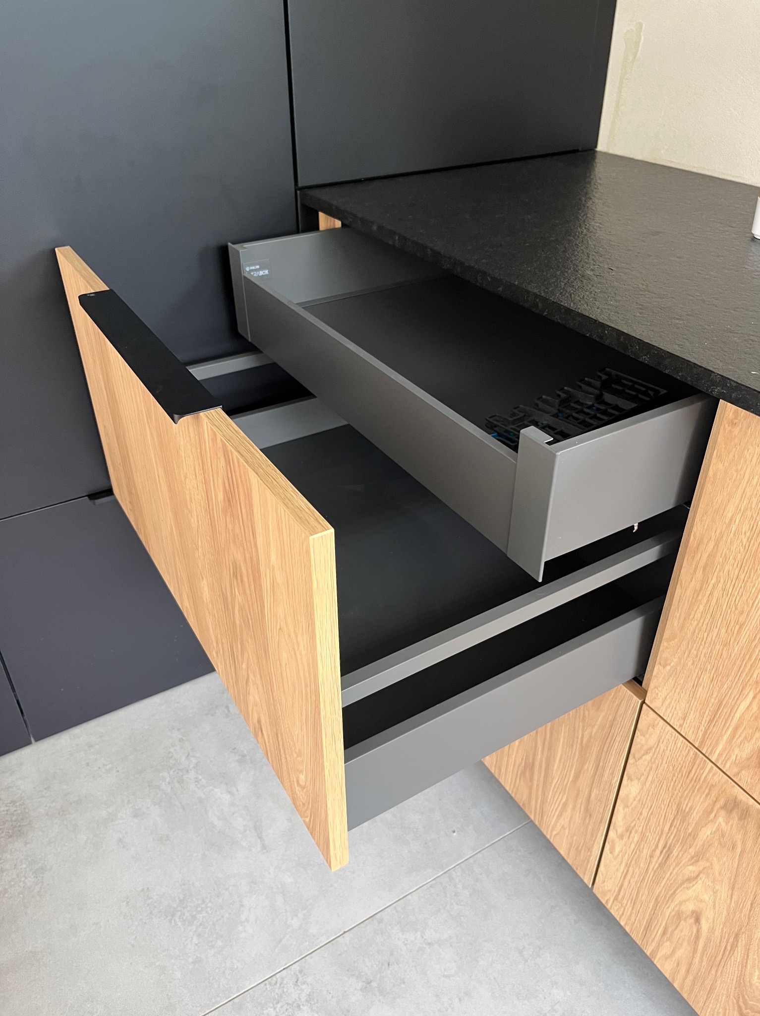 Otwarta szuflada z drewnianym frontem w nowoczesnej, grafitowej kuchni z czarnym blatem. Widoczne elementy szuflady i subtelne oświetlenie. Detal.