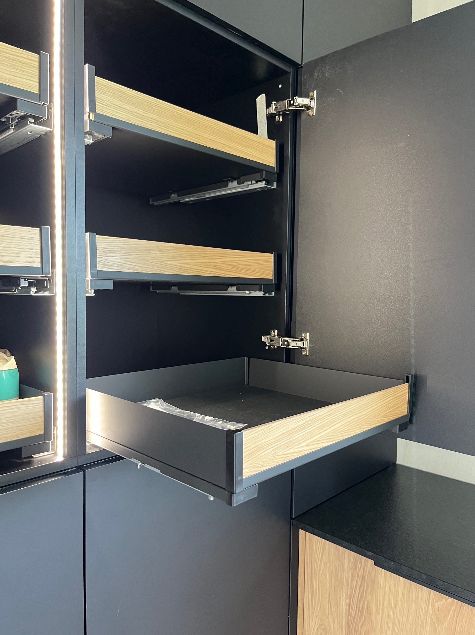 Ciemna szafa z wysuwanymi szufladami z jasnego drewna, widoczne prowadnice i okucia. Szuflady częściowo otwarte, wnętrze szafy oświetlone.