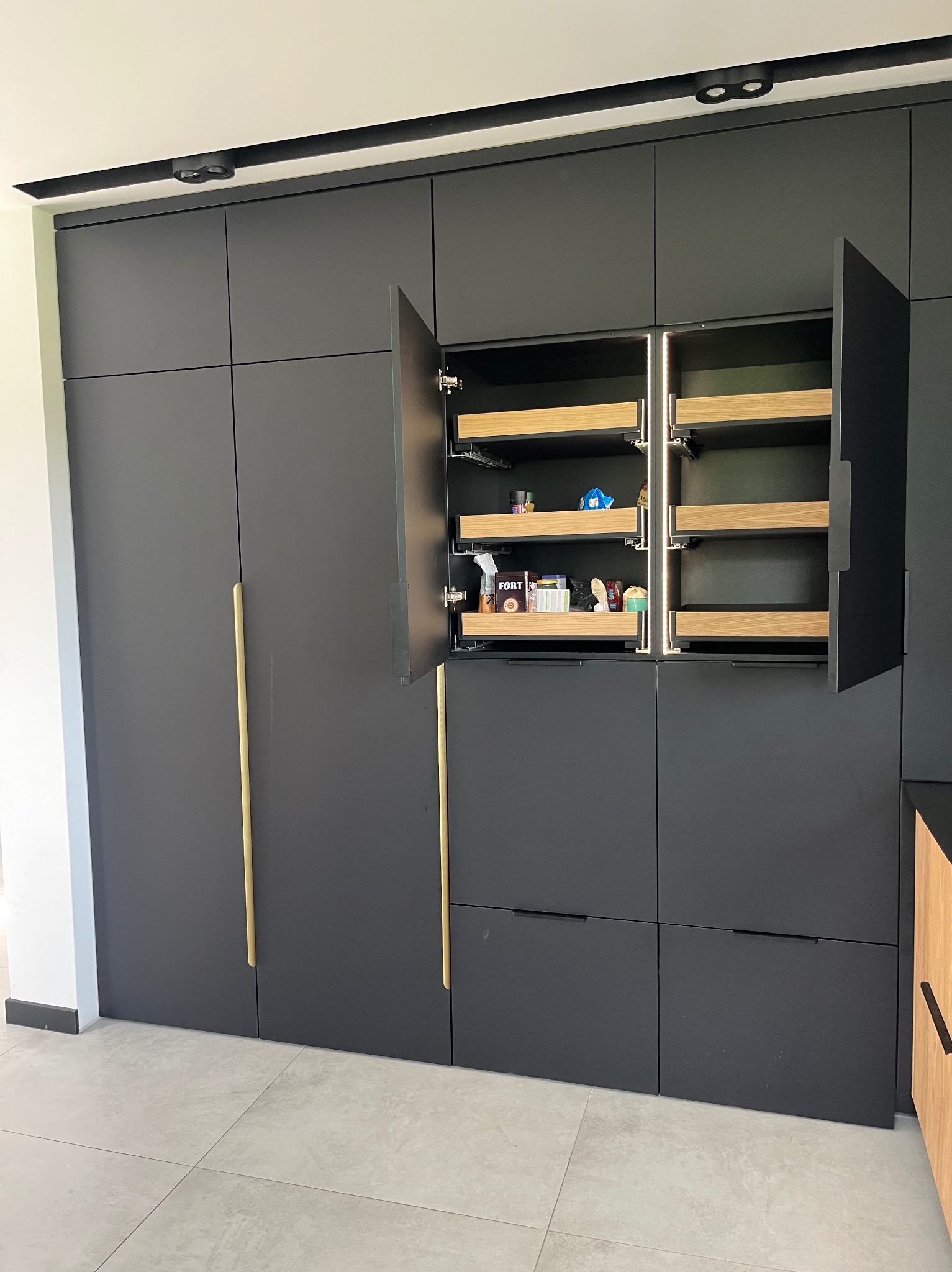 Nowoczesna zabudowa kuchenna w kolorze grafitowym z drewnianymi szufladami cargo, oświetleniem LED i złotymi uchwytami, na tle jasnej podłogi z płytek.