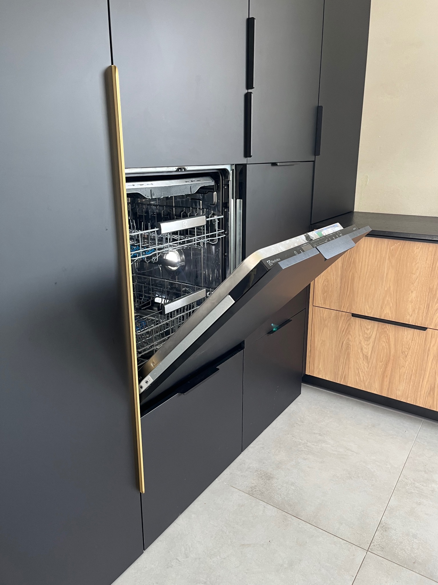 Nowoczesna kuchnia z szarymi szafkami i otwartą zmywarką Electrolux. Złote akcenty na frontach szafek. Widoczne wnętrze zmywarki i drewniane fronty sąsiednich szafek.