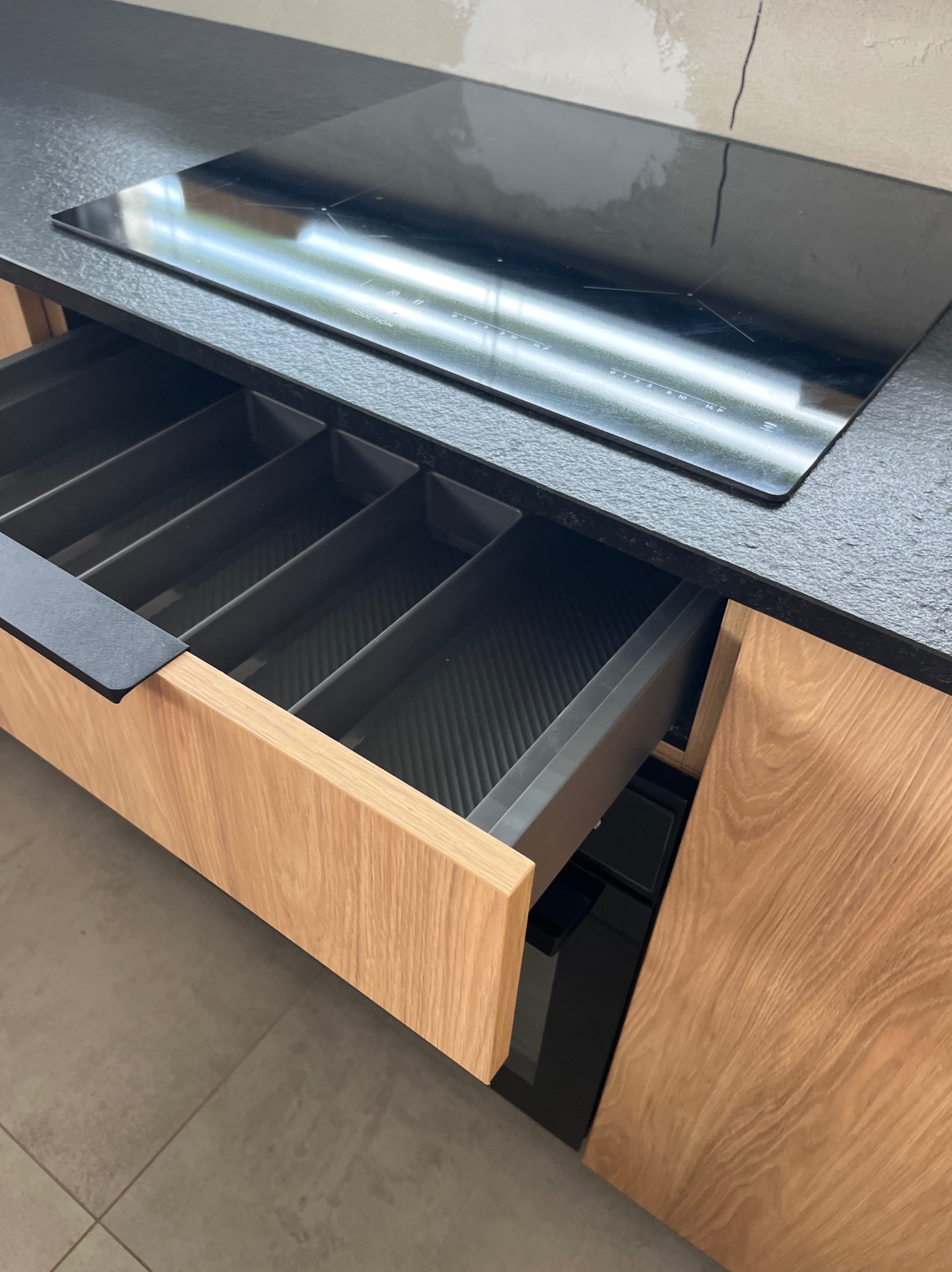 Otwarta szuflada z przegródkami w drewnianej zabudowie kuchennej, czarny blat z płytą indukcyjną. Minimalistyczny design, jasne drewno, szare płytki podłogowe.