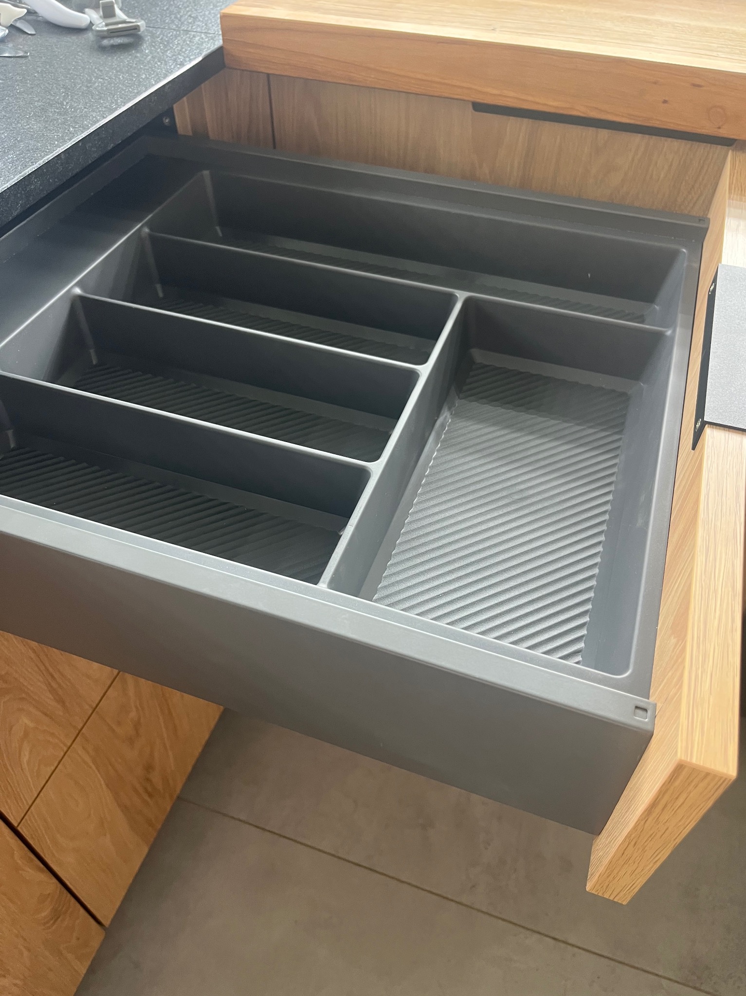 Otwarta szuflada z szarym organizerem na sztućce i akcesoria, wbudowana w drewnianą zabudowę kuchenną z blatem z ciemnego kamienia.