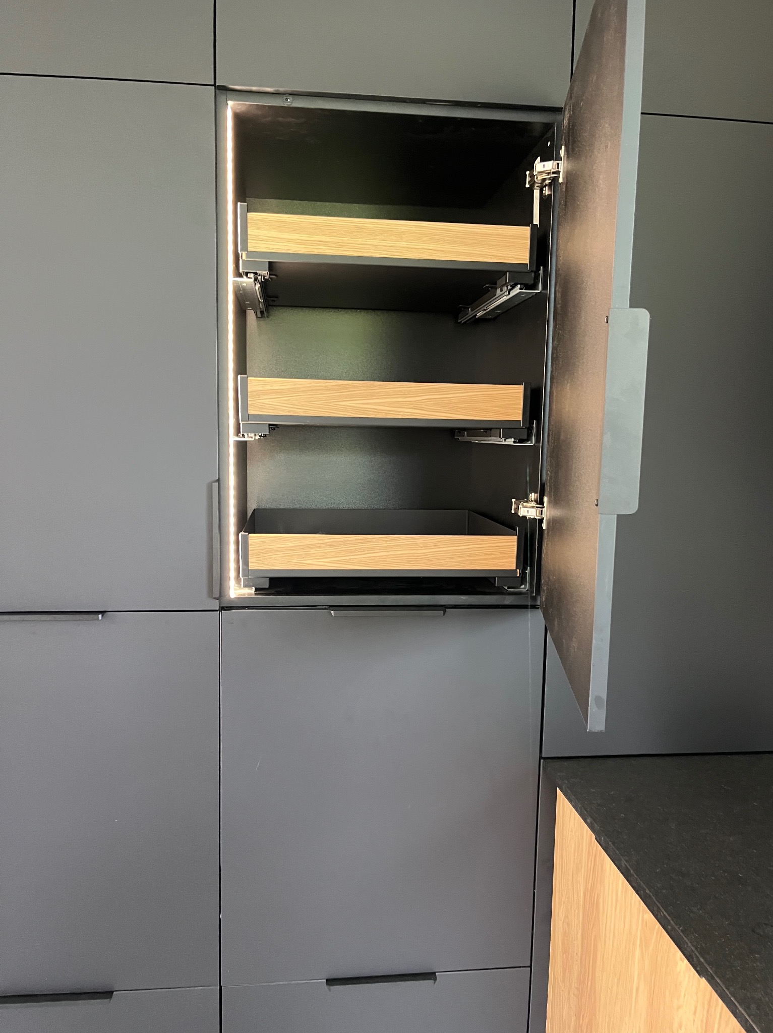 Szara szafa kuchenna z wewnętrznym oświetleniem LED, wysuwane półki z drewnianymi frontami i otwartymi drzwiczkami, widoczne zawiasy. Nowoczesny design.