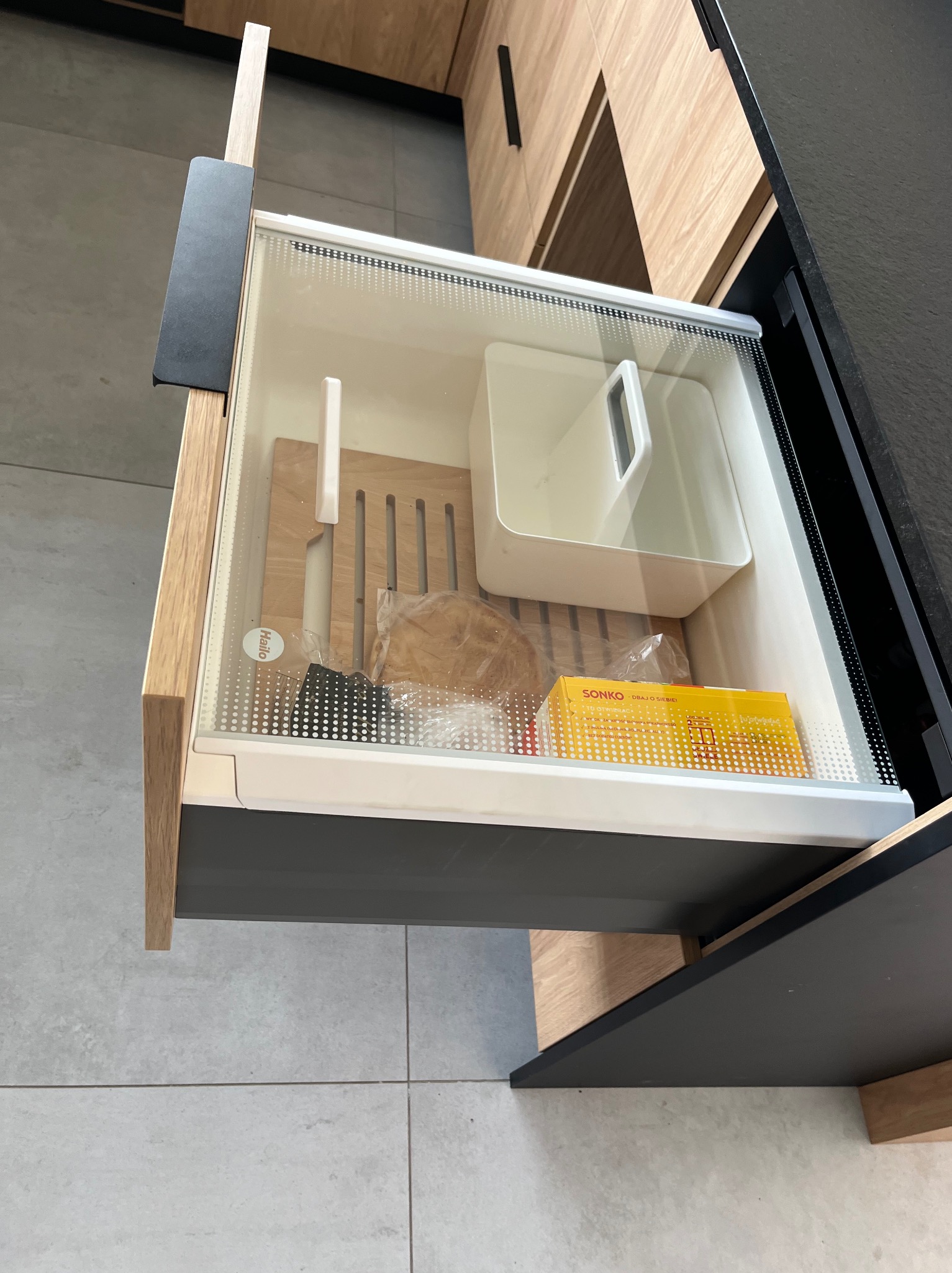 Otwarta szuflada kuchenna z przezroczystym frontem, ukazująca drewnianą deskę, pojemnik i pudełko. Jasne drewno i szary blat w nowoczesnej kuchni.