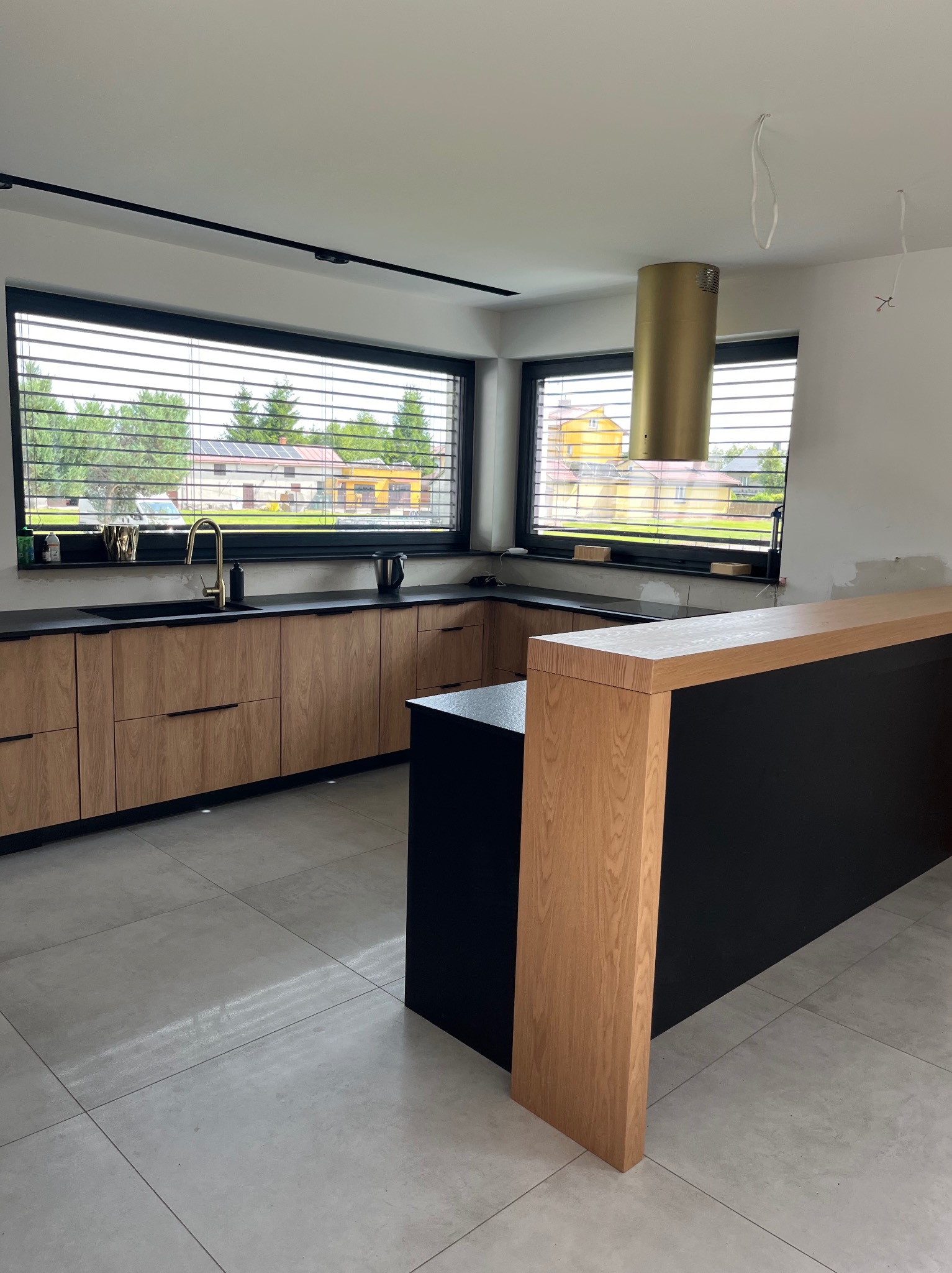 Jasna kuchnia z drewnianymi frontami i czarnym blatem, złota armatura i okap. Okno z żaluzjami. Elementy wykończeniowe.