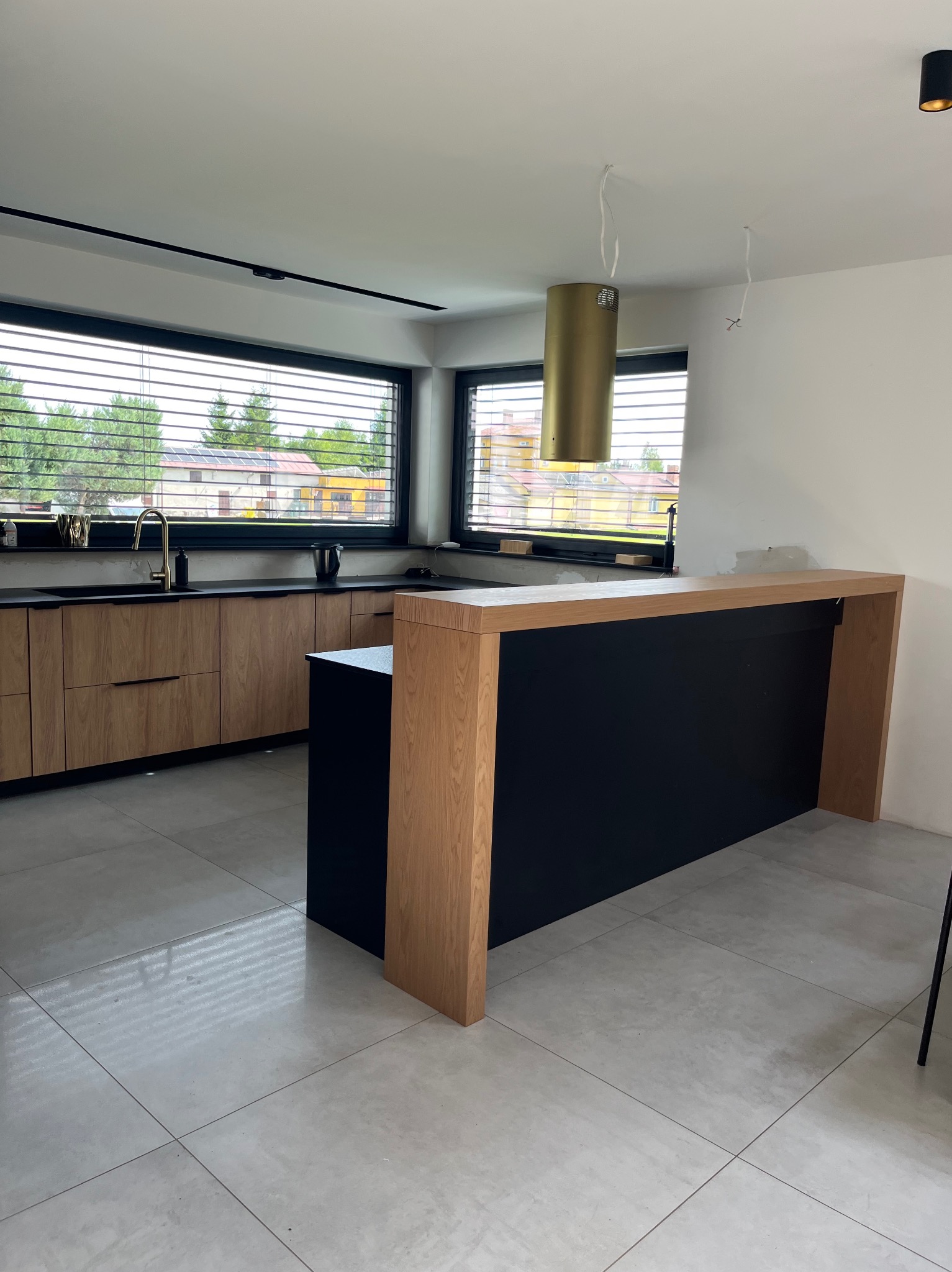 Nowoczesna kuchnia z wyspą: drewniane szafki, czarny blat, złota armatura i okap, szare płytki podłogowe. Duże okno z żaluzjami w tle.